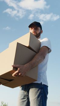 A deliveryman carrying cardboard boxes outdoors with a blue sky background, wearing a cap and casual clothes.