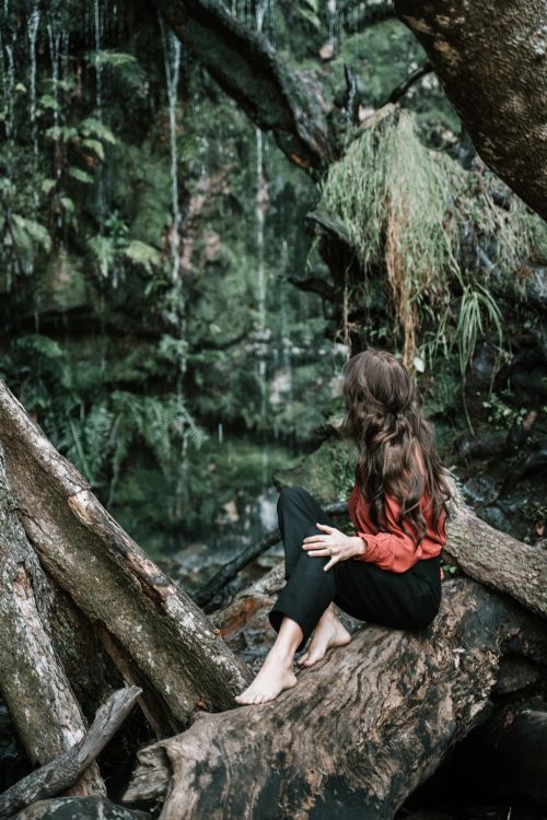 Free A woman sitting on a log by a forest waterfall, lost in thought. Stock Photo