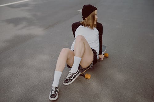 Woman in White and Black Shirt and Black Shorts Sitting on Her Longboard