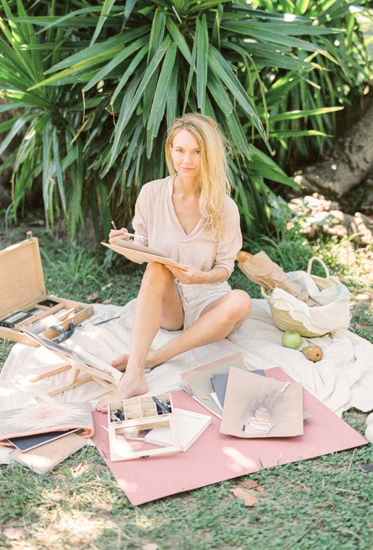 Blonde Woman Sitting On White Picnic Blanket Making A Sketch