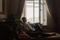 一名肌肉发达的男子穿着半敞的衬衫，懒洋洋地坐在窗前的躺椅上，神情轻松随意