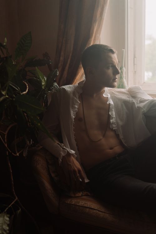 Free Subtle and elegant portrait of a shirtless man sitting by a window in a relaxed pose. Stock Photo