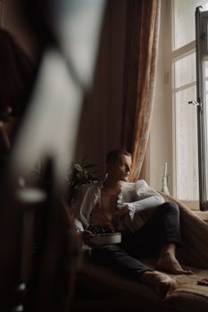 一名男子赤脚坐在窗前，端着一碗葡萄，在室内放松身心，散发着平静的心情