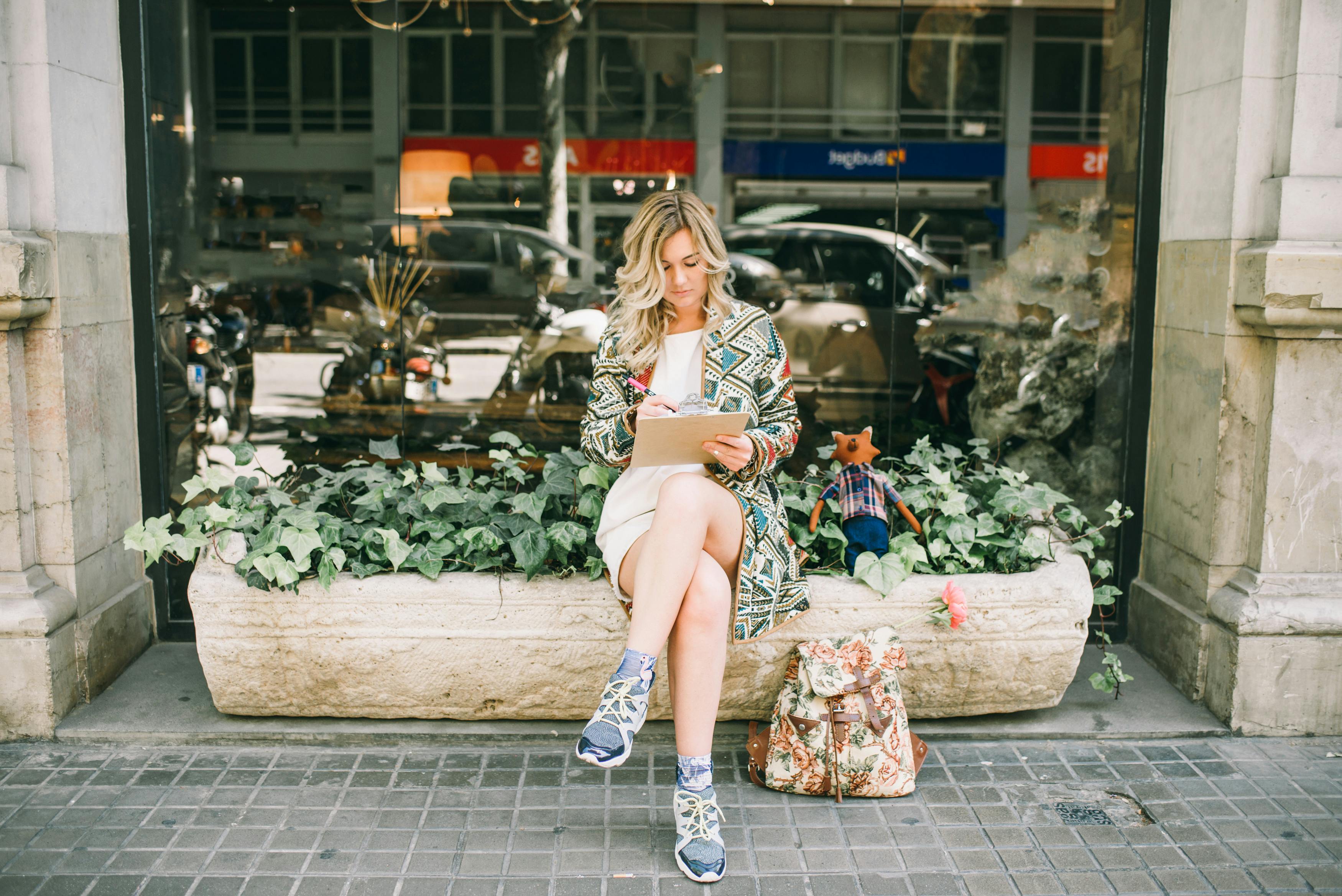 一位金发女子坐在花盆上，在写字板上写字，展现出时尚的现代风格