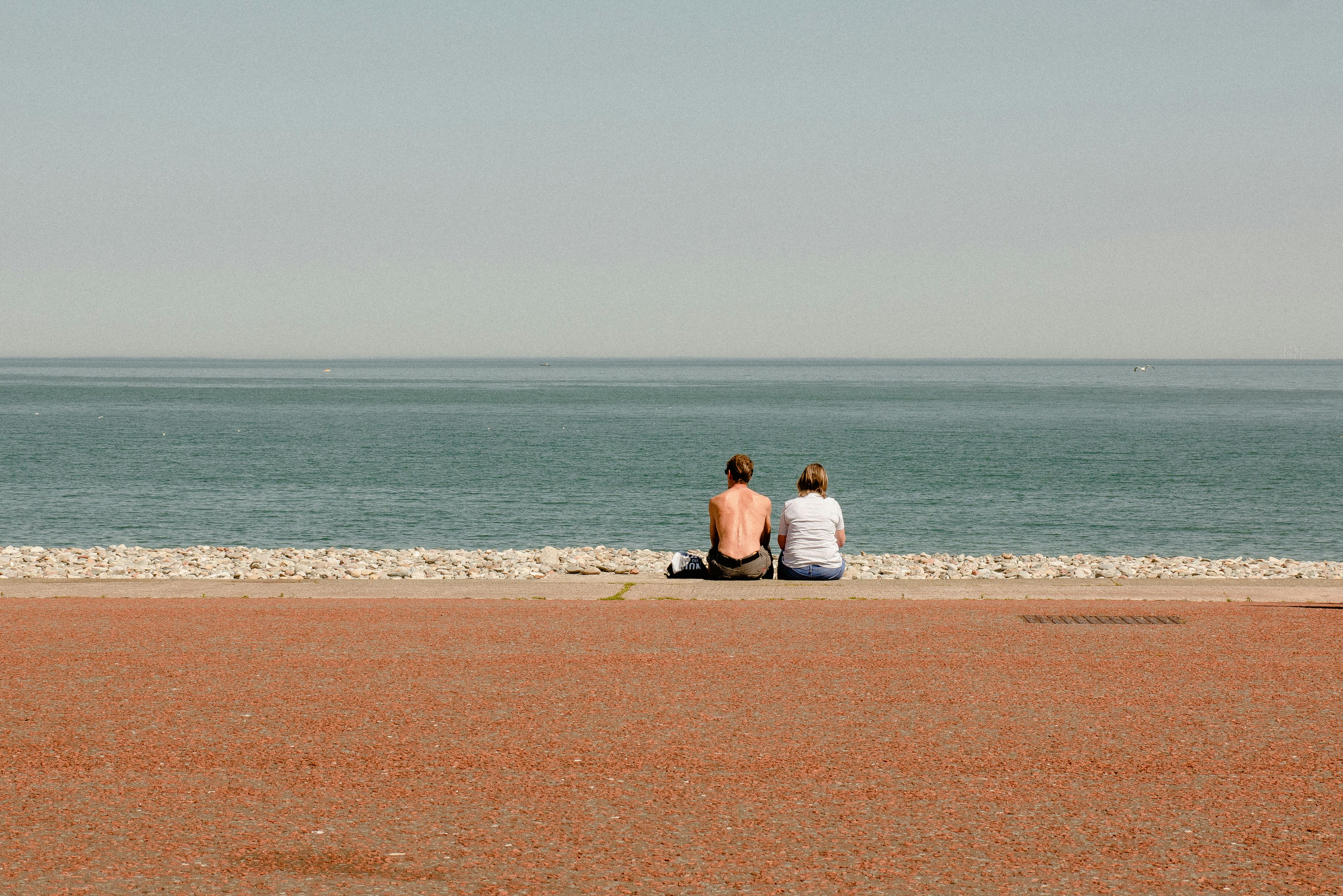 Back view of faceless couple sitting on stony coast of endless blue ocean in sunny weather