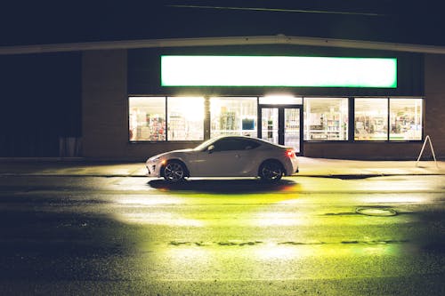 Free White coupe car on roadside at glowing shop with signboard late at night time Stock Photo