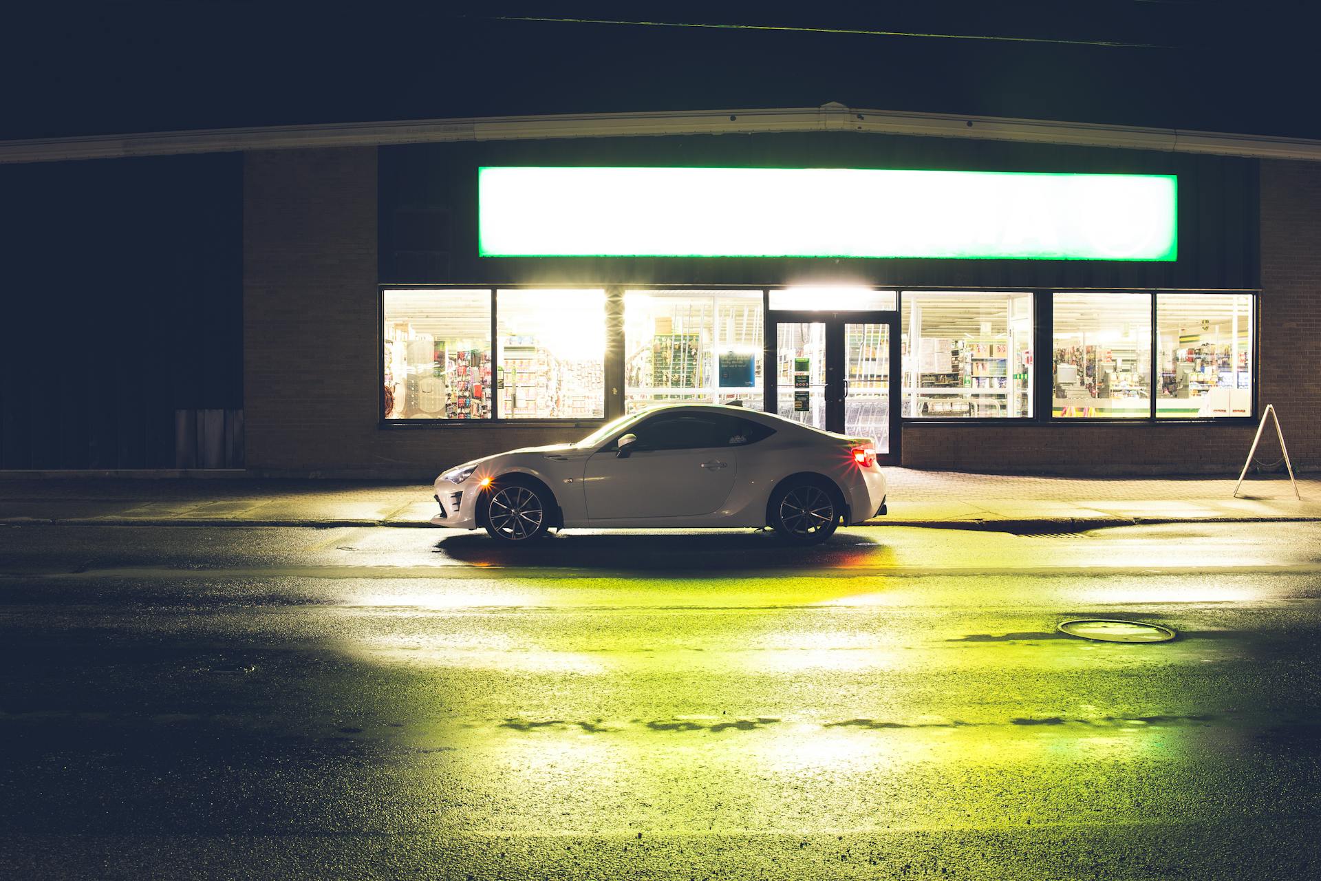 White coupe car on roadside at glowing shop with signboard late at night time