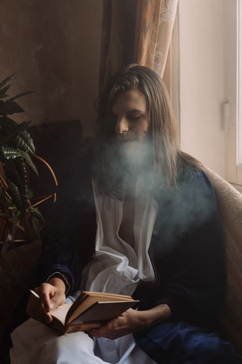 Free Androgynous person with long hair smoking and reading indoors in soft light. Stock Photo