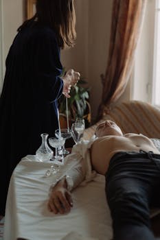 A shirtless man and a robed figure in a mysterious ritual scene with crystal glassware.