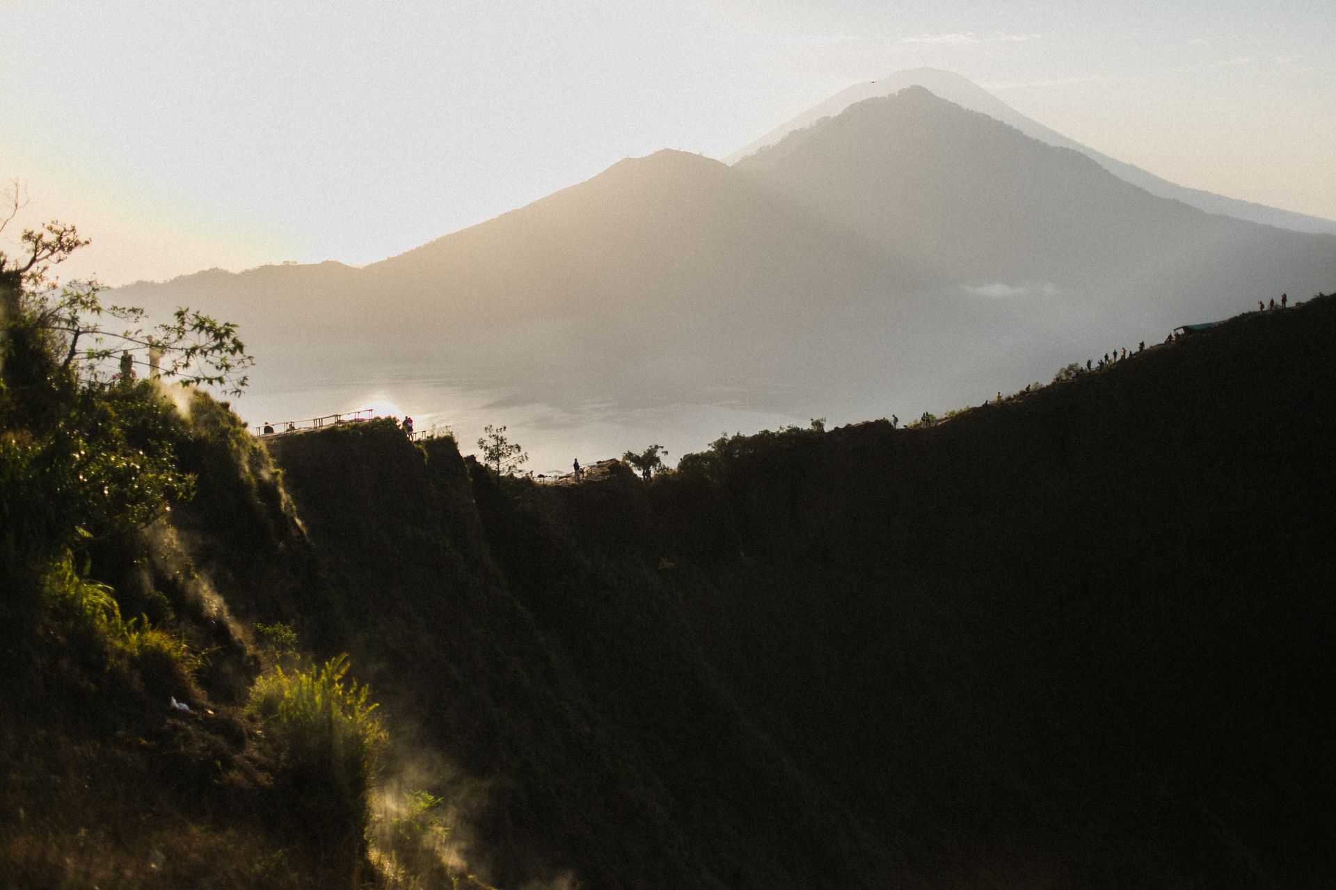 Green plants on steep rock and tall mountain ranges under light blue cloudless sky at sunset