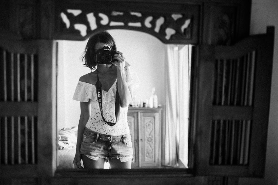 Free Black and white of casual female photographer in summer clothes standing against mirror in light room and taking photo with camera Stock Photo