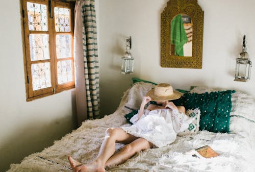 Anonymous woman in white dress covering face with straw hat while lying on bed in exotic hotel room