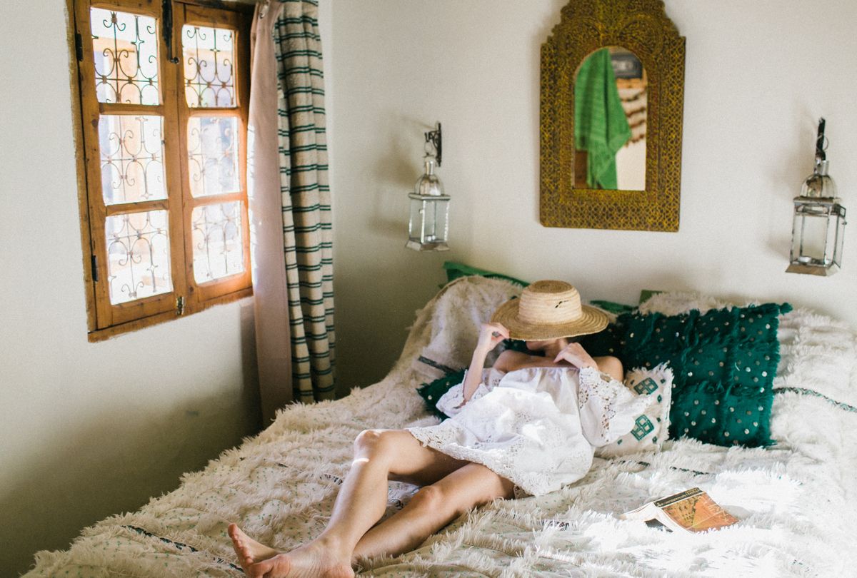 Anonymous woman in white dress covering face with straw hat while lying on bed in exotic hotel room Stock Photo.