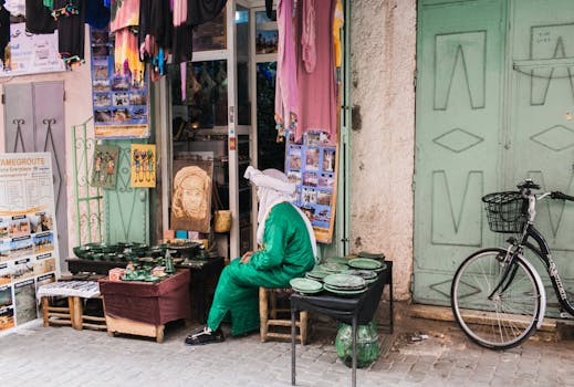 Local vendor in traditional attire seated outside a market shop with pottery and textiles.