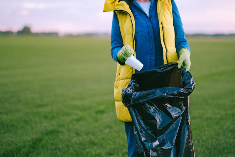 A Person Standing On Green Grass Field Putting Trash Inside The Garbage Bag