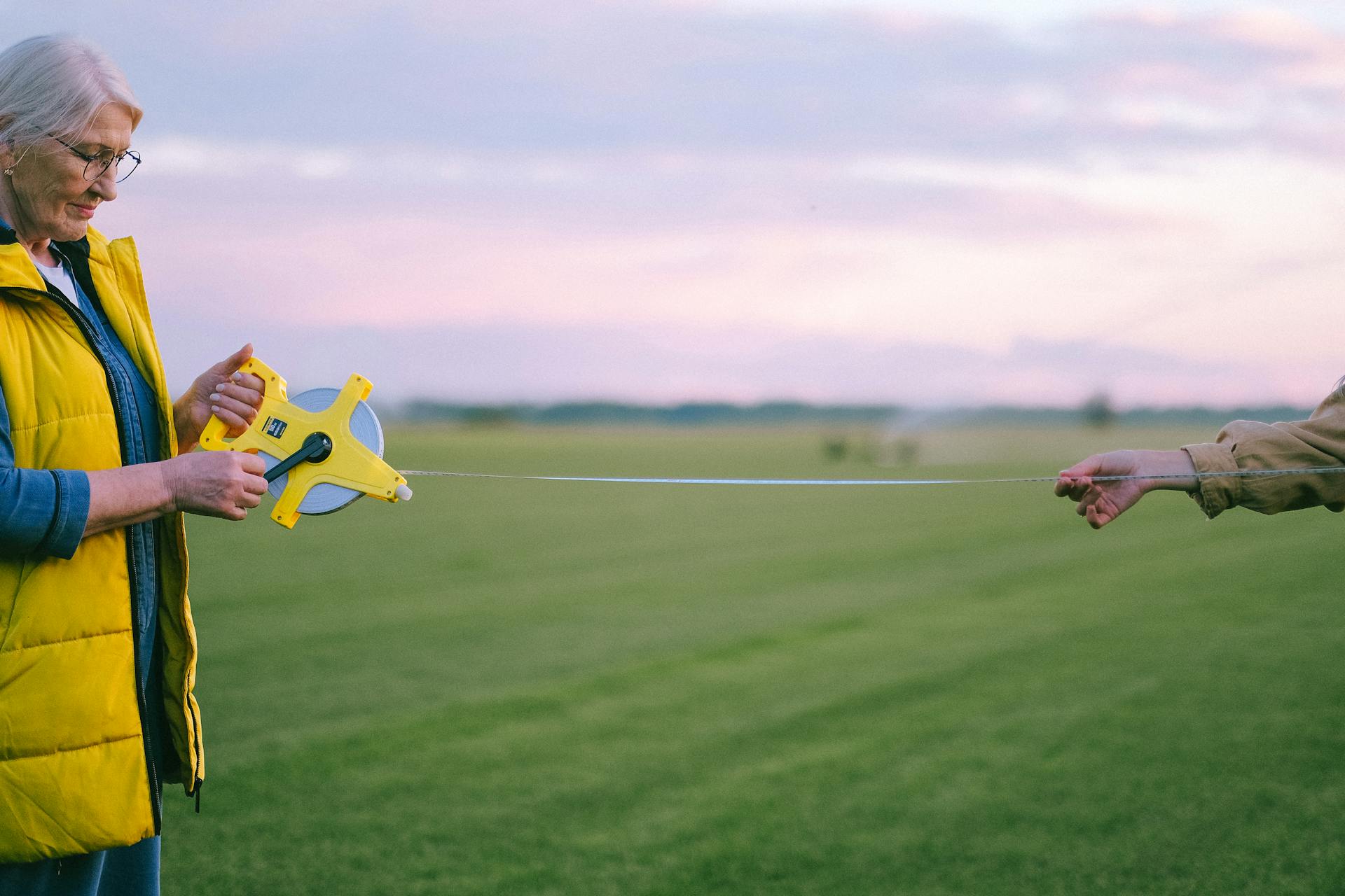 A senior woman uses a measuring tape on a grass field at sunset, showcasing agriculture tools and techniques.