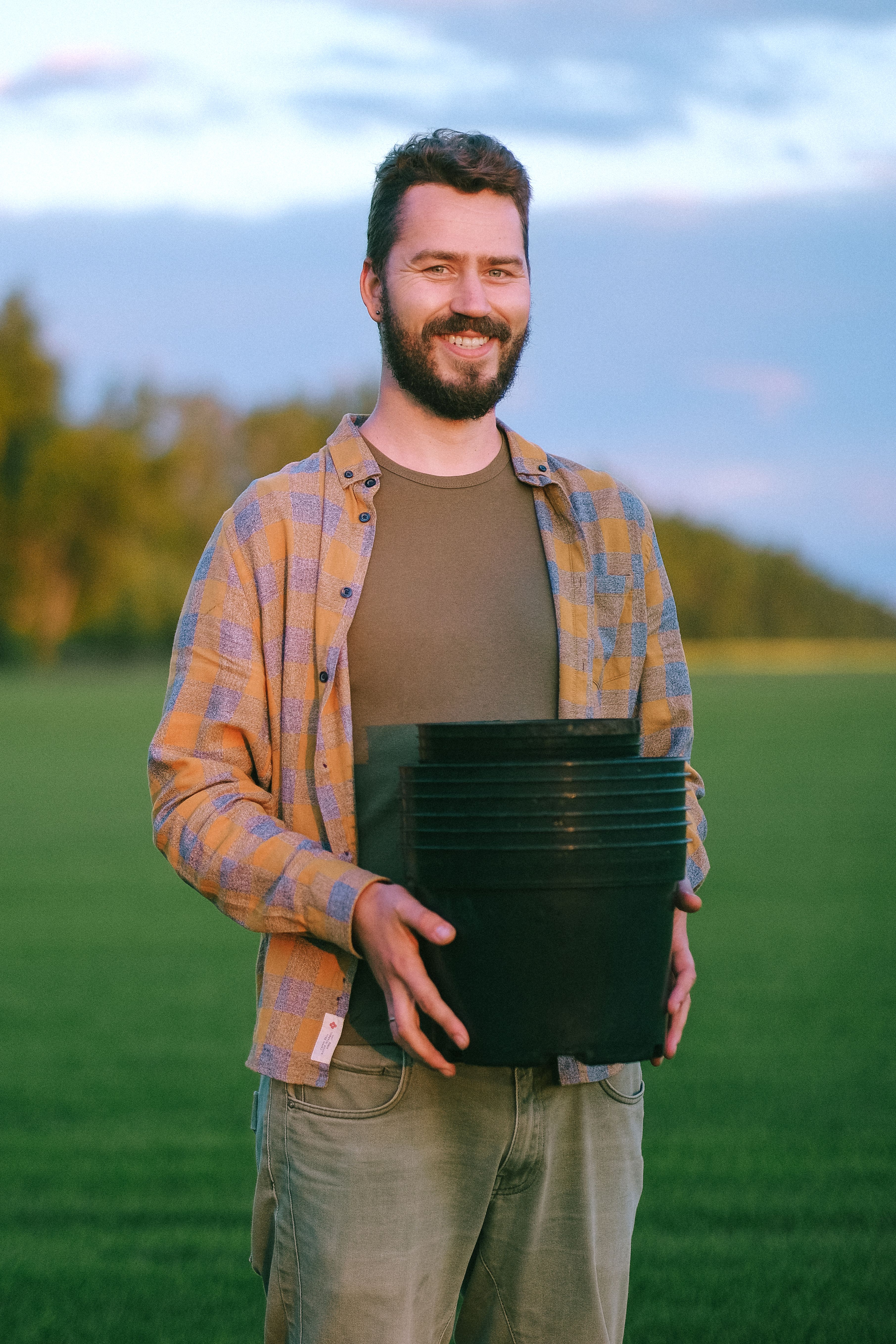 A smiling man holding a black pot in a green field during sunset, wearing a plaid jacket.