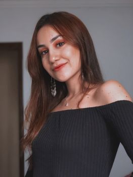 Portrait of a smiling young woman in an off-shoulder black top, exuding confidence and warmth indoors.