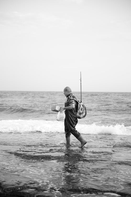 Grayscale Photo of a Fisherman Walking at the Beach