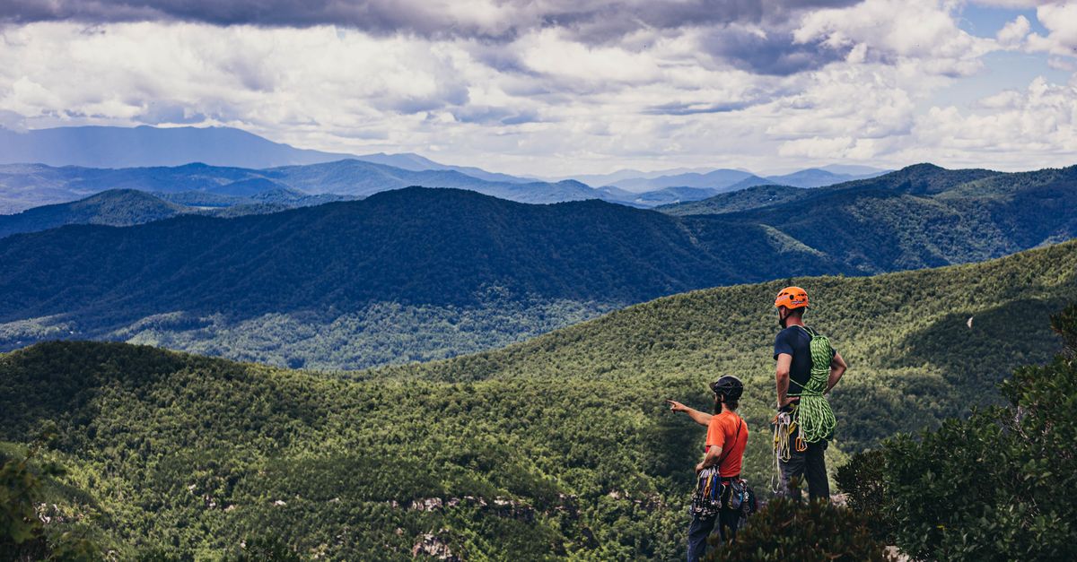 Two climbers with gear overlook lush green mountains under a cloudy sky.