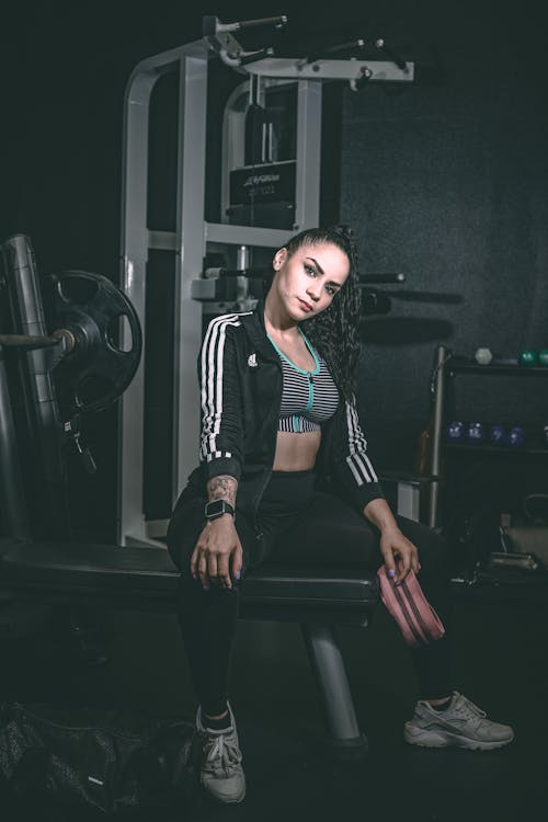 Young attentive fit female athlete in sportswear sitting on exercise equipment while looking at camera in gymnasium
