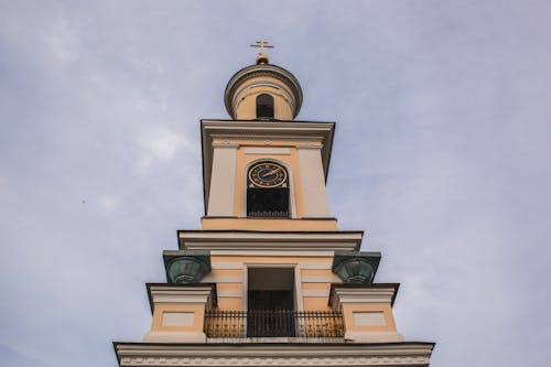 From below of aged church tower with golden cross and rounded clock over balcony in cloudy day