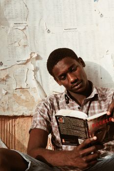 Concentrated African American male in checkered shirt leaning on shabby wall with board with papers while reading book in daylight