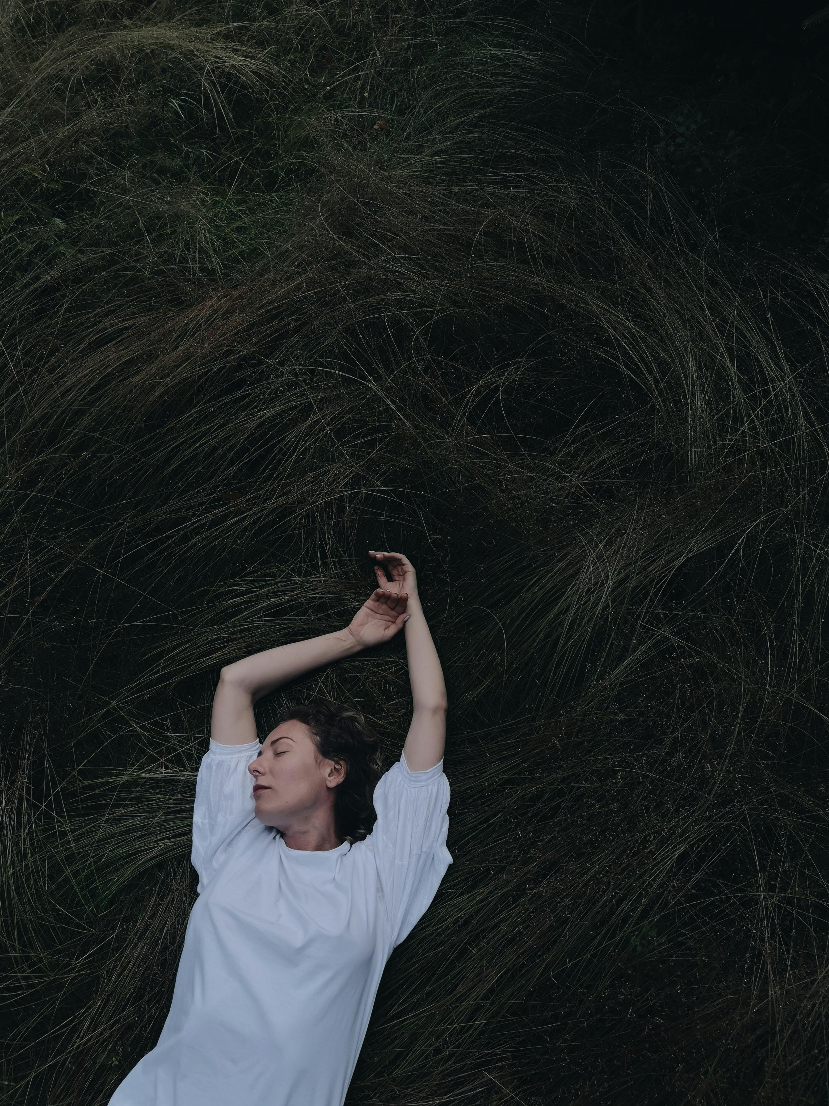 A serene woman in a white top lies with eyes closed on a grassy field, embracing calmness.