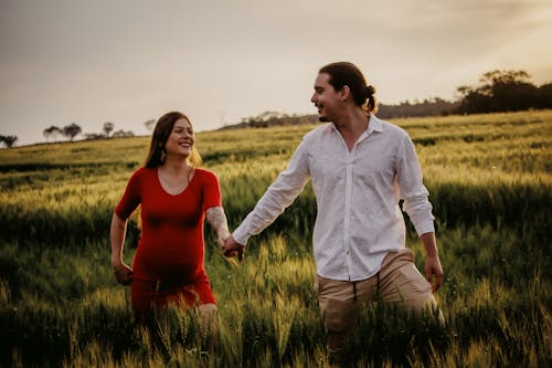 Pregnant Woman and Man Looking at Each Other While Holding Hands on Grass Field 