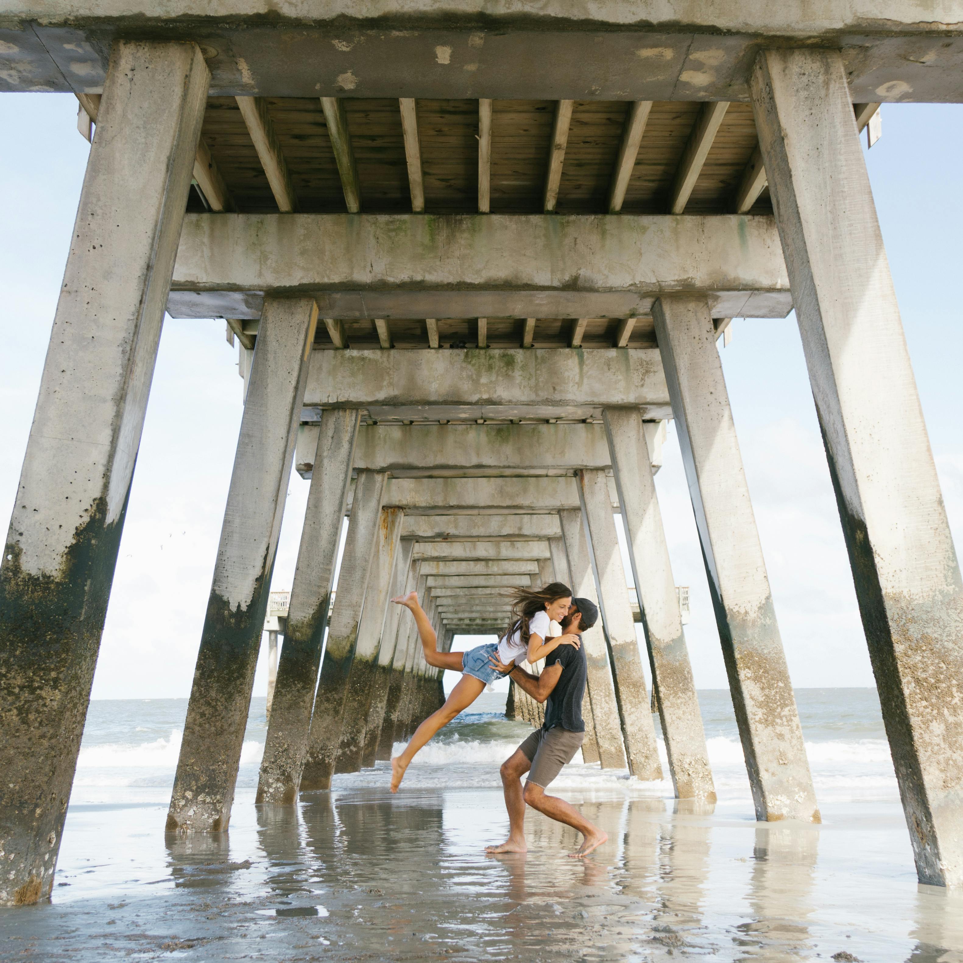 Couple embraces romantically under a seaside pier with waves in the background.