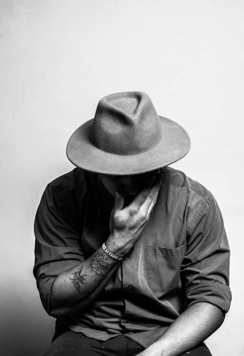 Black and white of anonymous young tattooed male in trendy outfit and hat touching face and looking down while sitting in studio