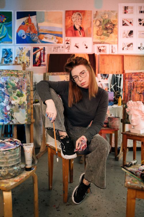 Free A female artist sitting in a colorful studio surrounded by her paintings, showcasing creativity and style. Stock Photo