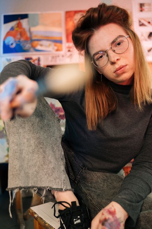 Free Creative female artist sitting in a vibrant workshop, holding a paintbrush with expression and focus. Stock Photo