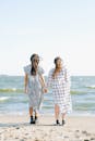 两位亲密女性身着连衣裙，手牵手站在海边沙滩上的背影