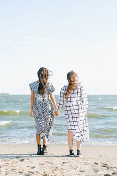 两位亲密女性身着连衣裙，手牵手站在海边沙滩上的背影
