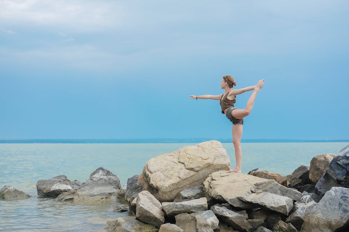 Full body of young concentrated female in comfortable wear doing yoga on rocky coast of sea