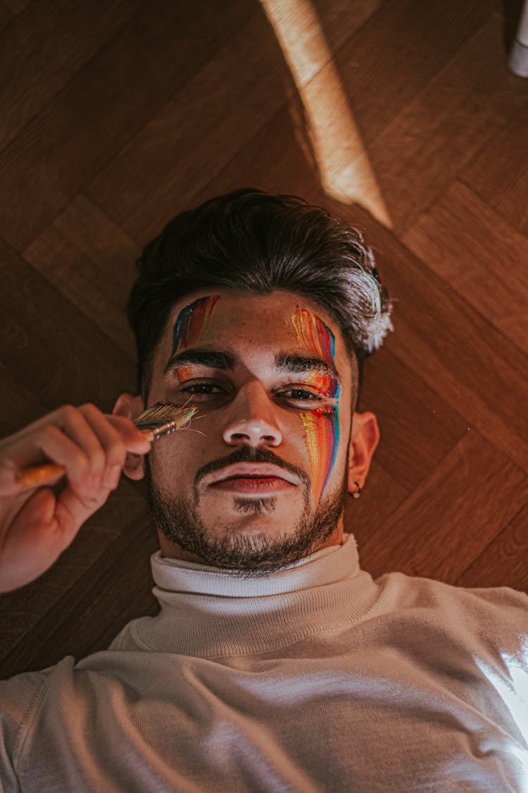 Man Lying On Floor And Painting Multicolored Lines On Face