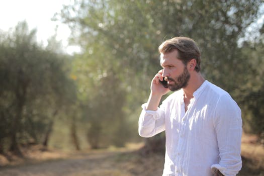 Bearded man in white shirt talking on phone in a sunny outdoor setting with blurred background.