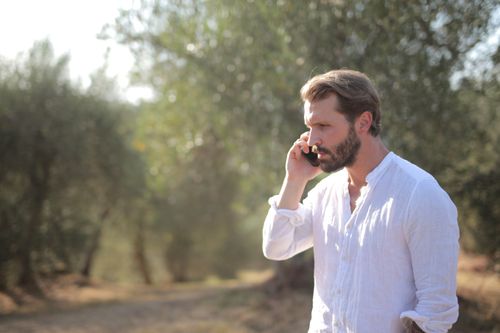 Free Bearded man in white shirt talking on phone in a sunny outdoor setting with blurred background. Stock Photo
