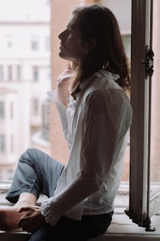 A sophisticated young man in a white shirt poses by the window, embracing natural light.