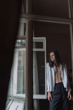 一位成熟的雌雄同体男性在公寓窗边摆出姿势，散发着优雅与风格