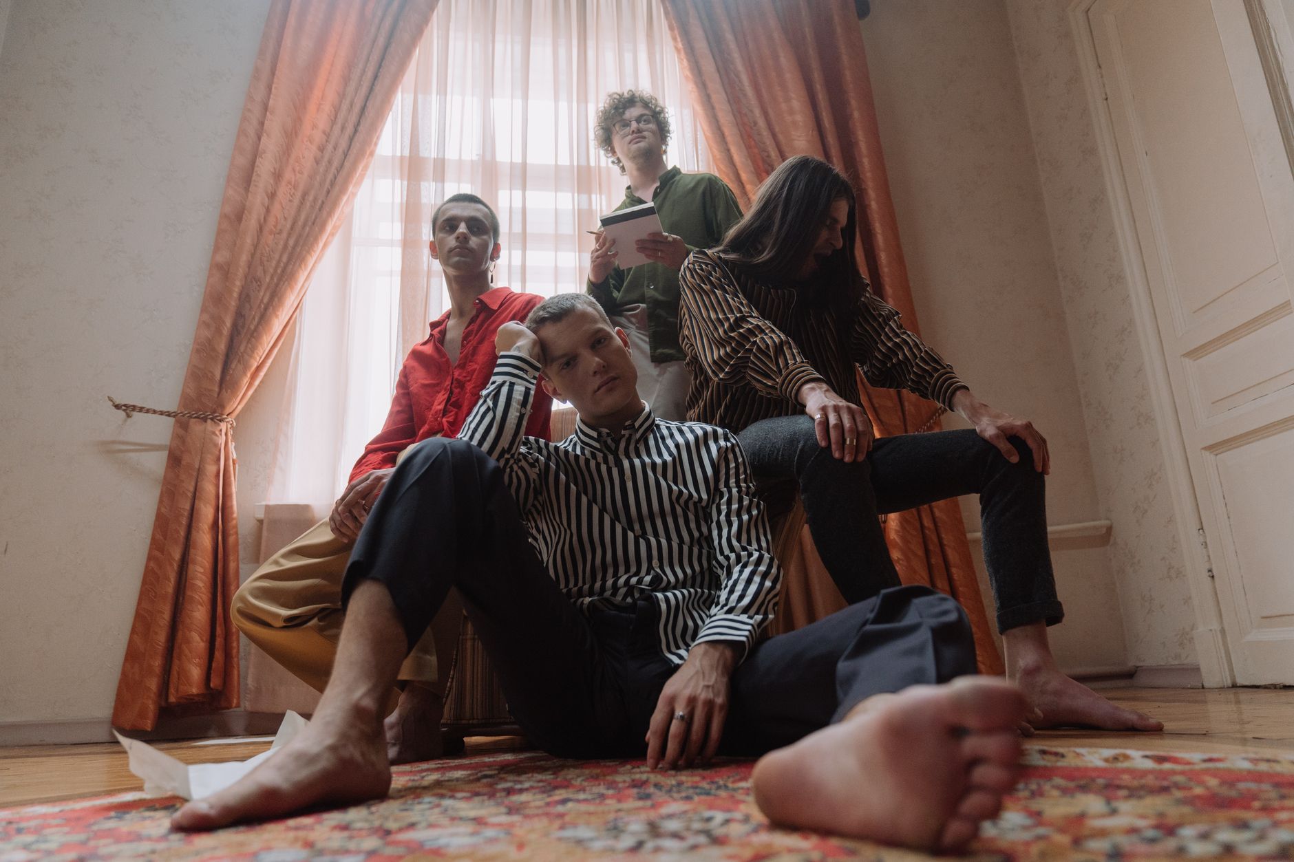 A cinematic group photo of four diverse barefoot men posing indoors with dramatic curtains.
