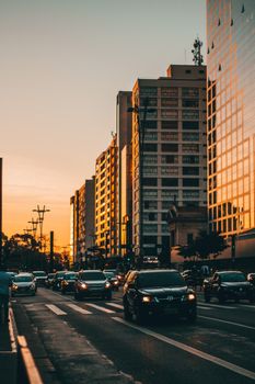 柏油路上行驶着各种汽车，映衬着风景如画的日落天空，倒映在现代多层建筑的玻璃镜立面上