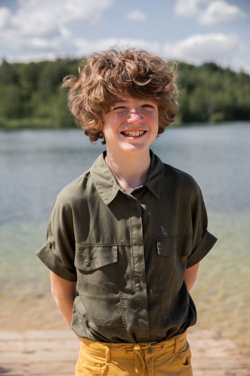Free A cheerful child with curly hair and a green shirt smiling by a scenic lake on a sunny day. Stock Photo