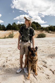 A teenager standing with a German Shepherd dog on a sandy outdoor landscape under a blue sky.