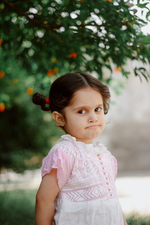 Free A young girl stands under a fruit tree, wearing a pink and white dress, expressing a thoughtful expression. Stock Photo