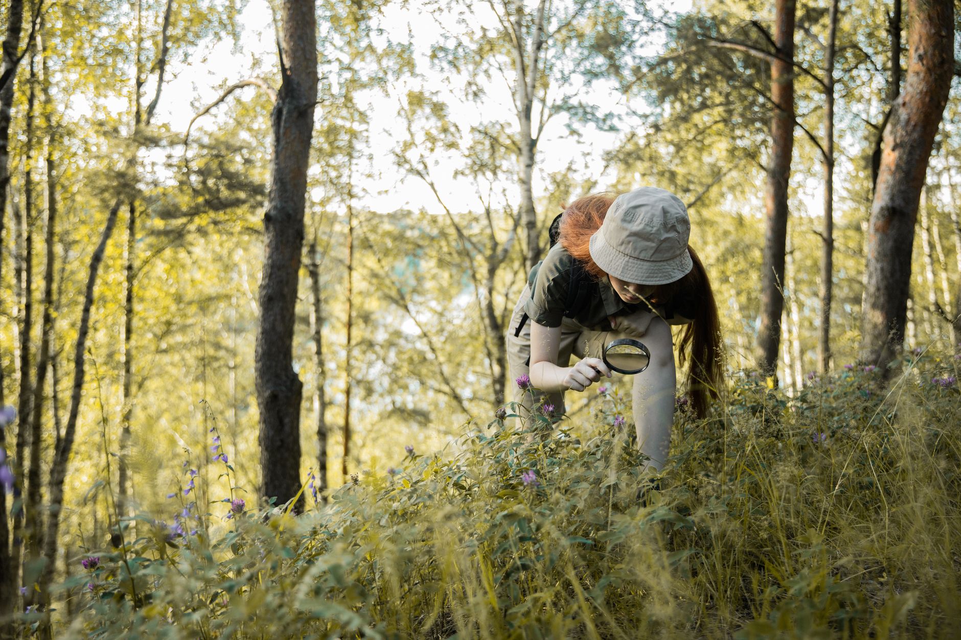 A redhead teenager with a hat investigates wildflowers in a forest with a magnifying glass.