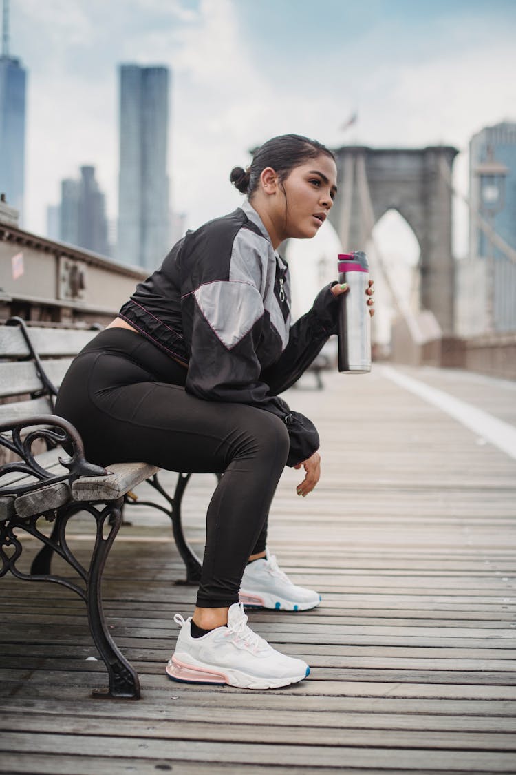 Woman In Sports Clothing Sitting On A Bench And Drinking Water 