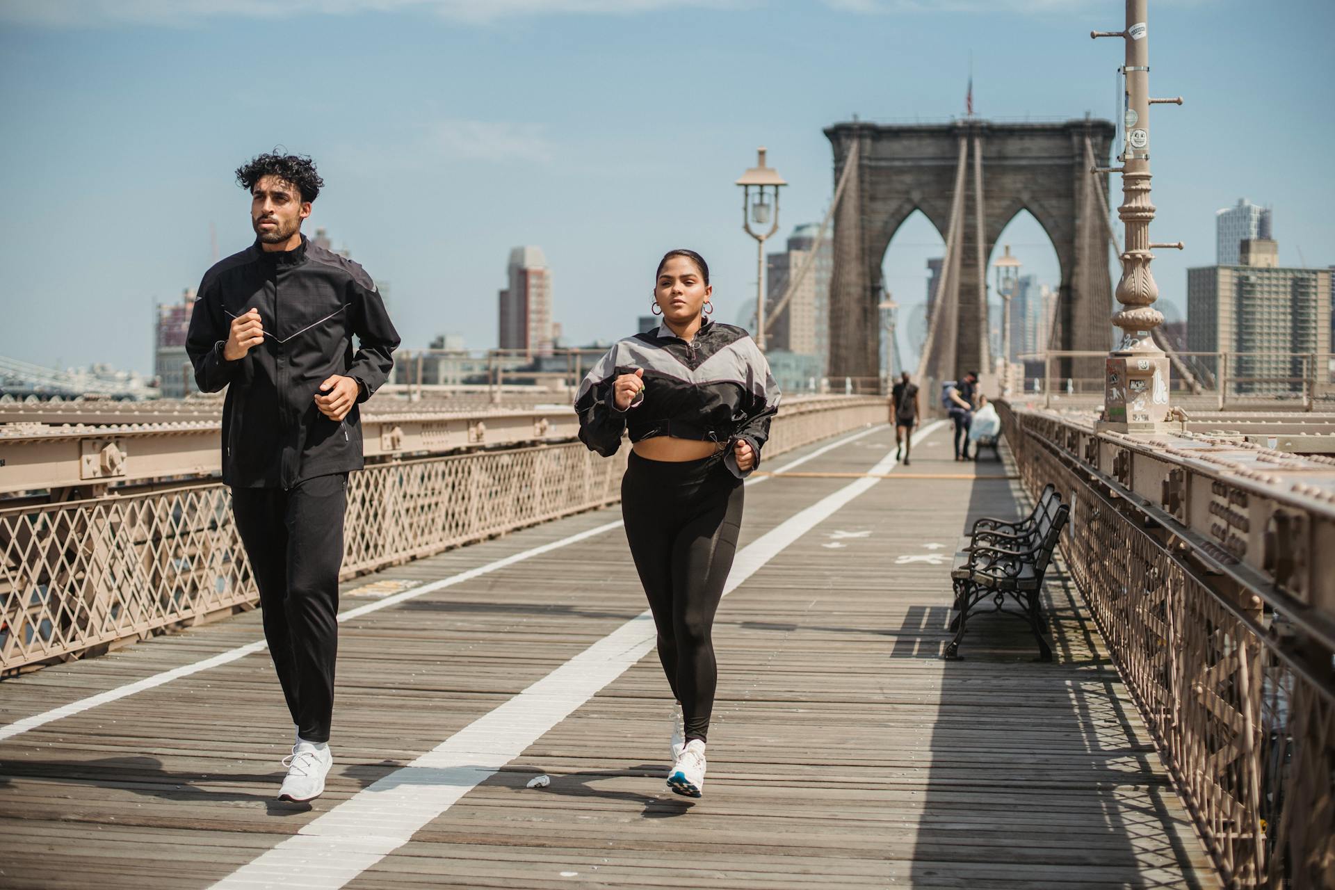 Two adults jogging on Brooklyn Bridge with city skyline in the background, showcasing health and vitality.