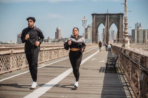 Free Two adults jogging on Brooklyn Bridge with city skyline in the background, showcasing health and vitality. Stock Photo